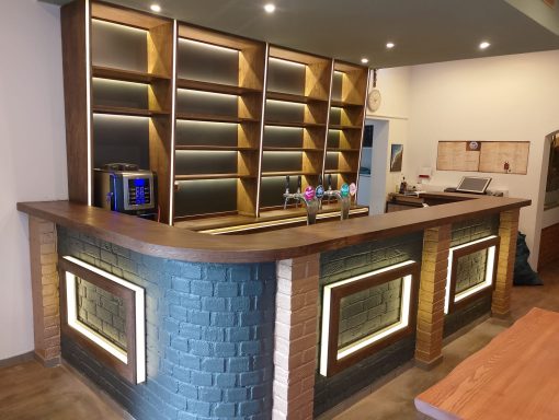 Moderne Bar mit Holztheke, beleuchtetem Regal und Mikrowelle im Hintergrund.