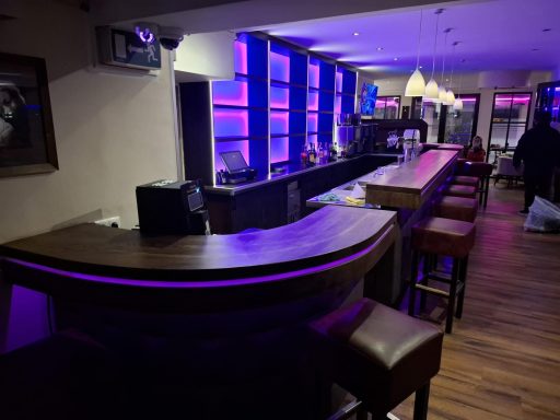 Moderne Bar mit Holztheke und lila Beleuchtung, Stühle entlang der Theke.