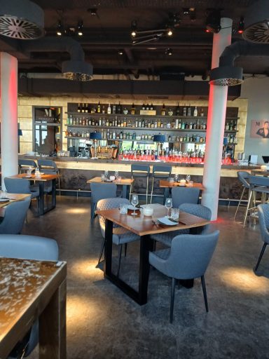 Moderne Restaurantansicht mit Holztischen, Stühlen und einer Bar im Hintergrund.