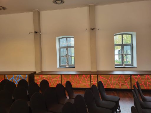 Raum mit zwei Fenstern, bunten Wandverkleidungen und Stuhlreihen.