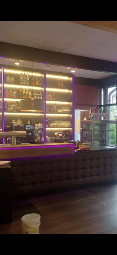 Moderne Bar mit lila Beleuchtung und Holzregalen, Sitzbereich im Vordergrund.