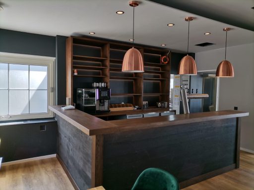 Moderne Café-Bar mit Holztheke und Kupferlampen, hellen Wänden und Fenster.
