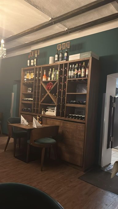 Holzregal mit Weinflaschen und Gläsern, daneben Tisch mit Stühlen in einem Restaurant.