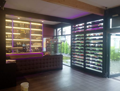 Moderne Bar mit lila Beleuchtung, Holzregal und Blick auf eine grüne Terrasse.