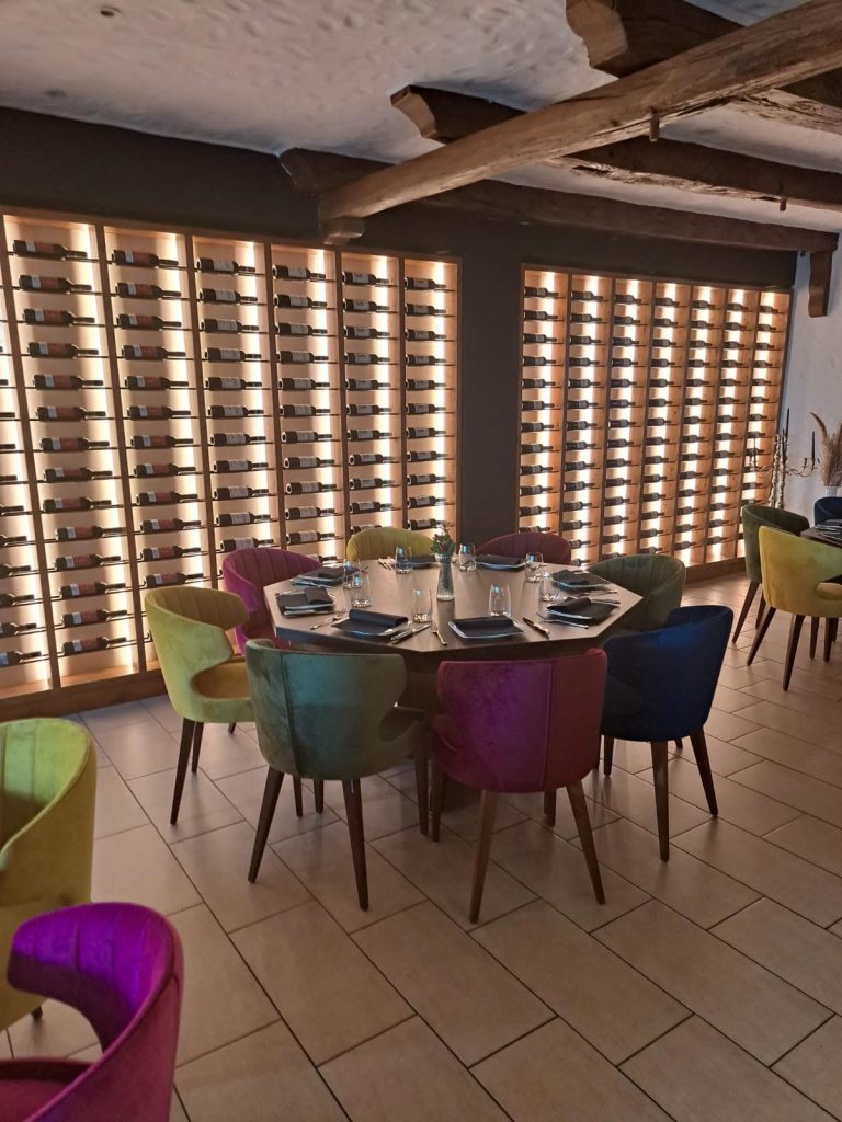 Bunte Stühle um einen Tisch vor einer Wand mit Weinfächern beleuchtet.