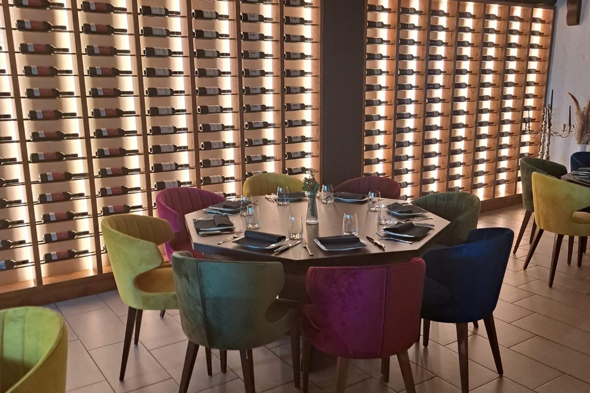 Bunte Stühle um einen runden Tisch, umgeben von einer Wand mit Weinregalen.
