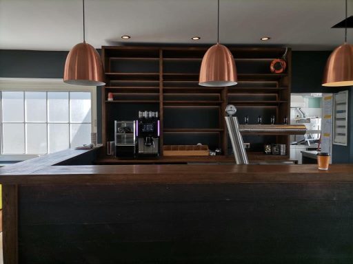 Moderne Café-Bar mit Holztheke und Kupferlampen, offene Regalwand im Hintergrund.