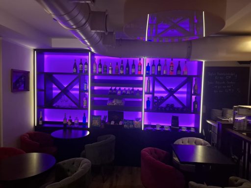 Moderne Bar mit violett beleuchtetem Regal und runden Tischen in entspannter Atmosphäre.